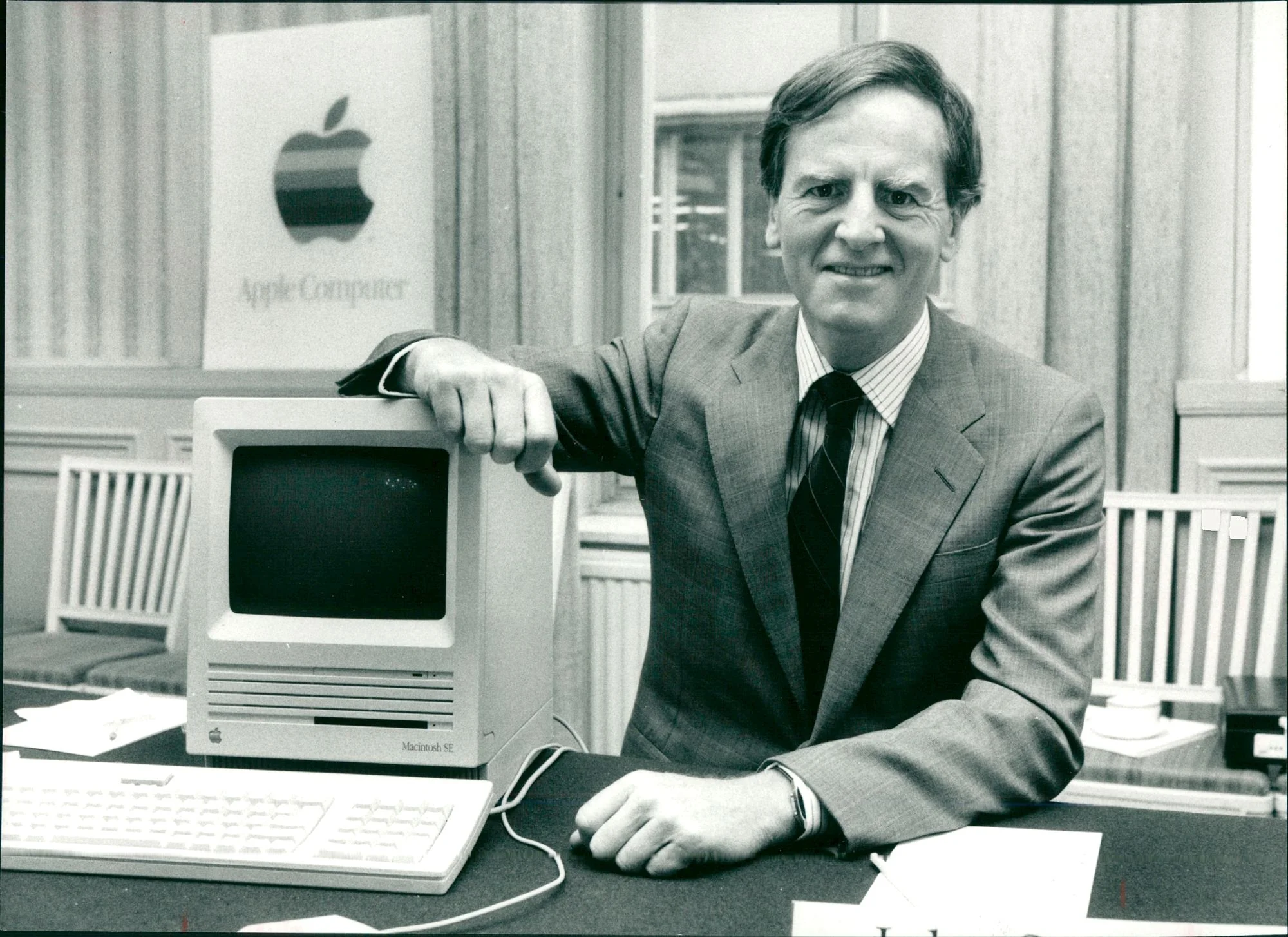 John Sculley smiles infront of a vintage Apple Computer and Apple logo.